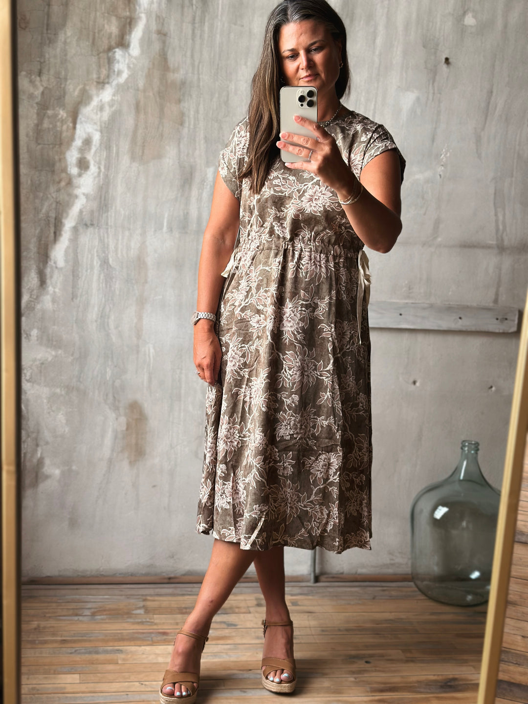 Woman taking a mirror selfie wearing a patterned dress in a room with wooden floor and concrete wall.
