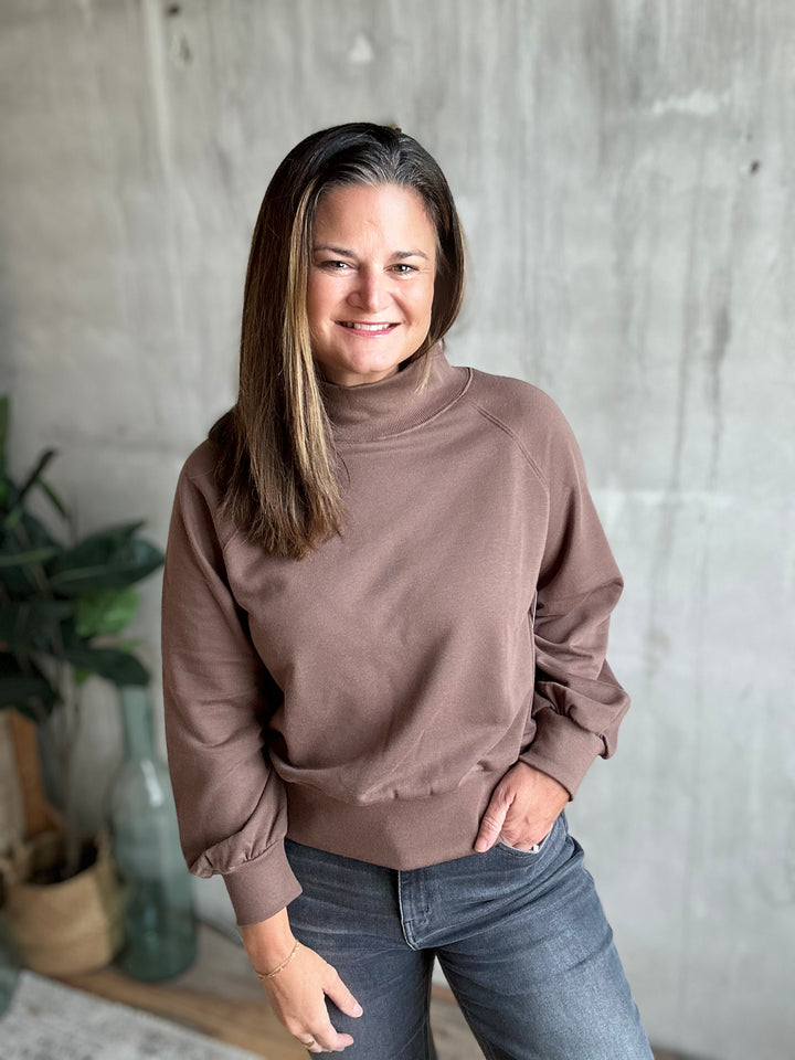 A woman with long brown hair stands indoors wearing a taupe brown mock neck long-sleeve sweatshirt and dark gray jeans, posing in front of a textured concrete wall.