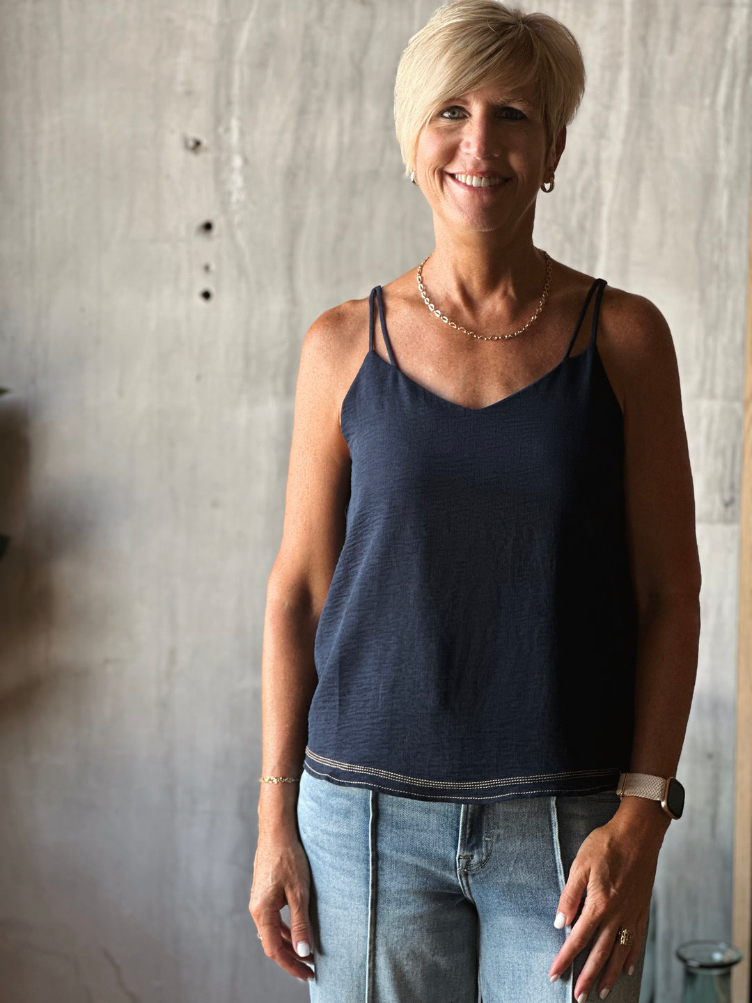 Woman wearing a navy blue tank top and light blue jeans standing against a textured wall.