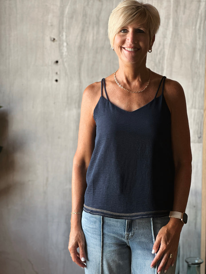Woman wearing a navy blue tank top and light blue jeans standing against a textured wall.