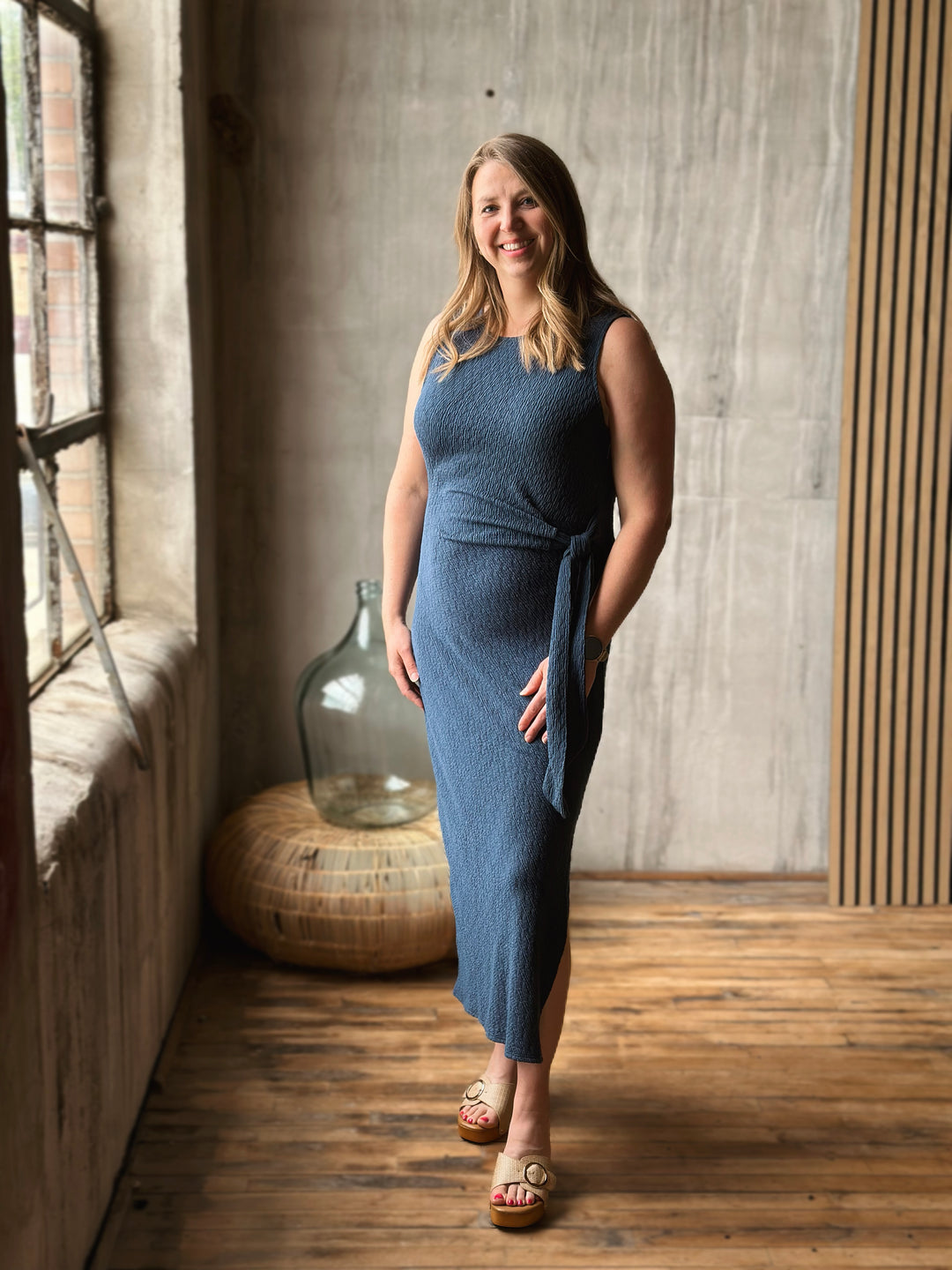 Woman in a blue dress with a knot standing in a room with a wooden floor and large window.