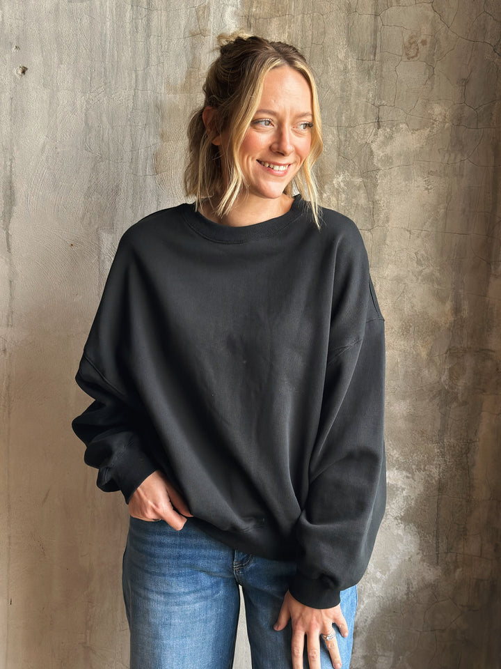 Woman wearing a black sweatshirt and blue jeans against a textured wall.