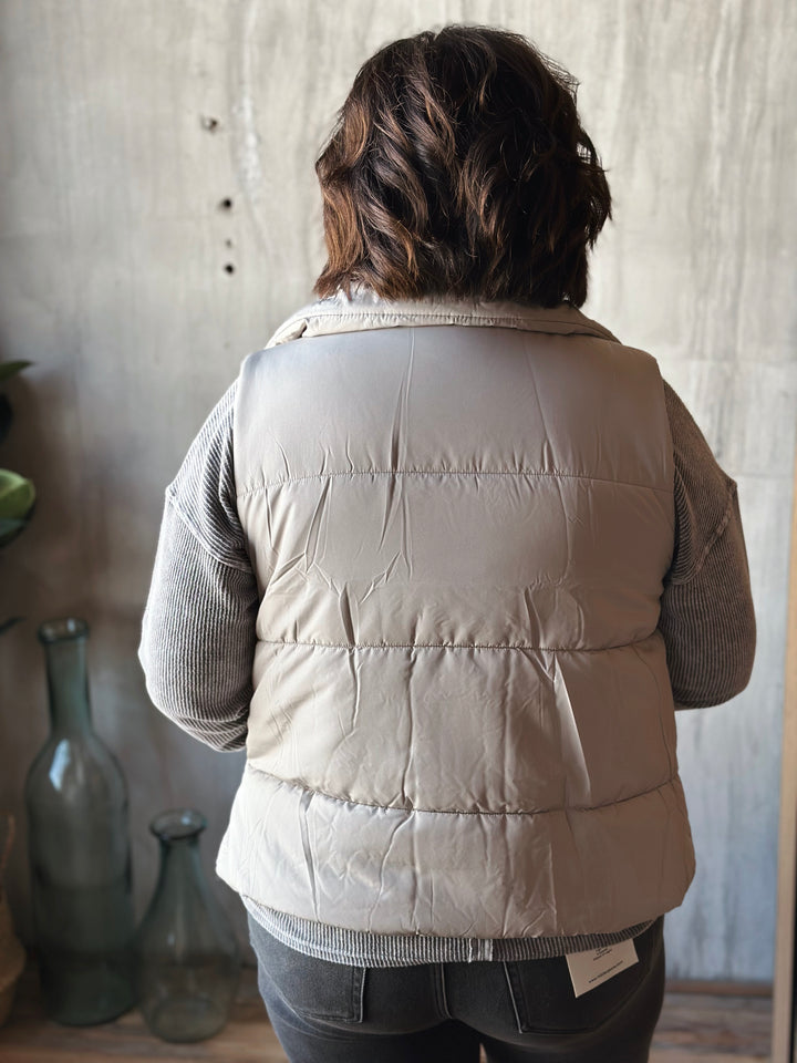 Person wearing a beige puffer jacket standing in a room with a textured wall and bottles.