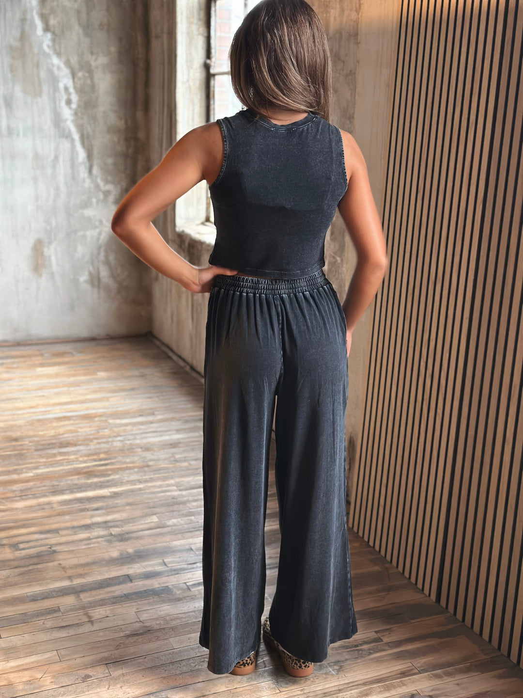 Person wearing a black sleeveless top and wide-leg pants standing in a room with wooden flooring and a textured wall.