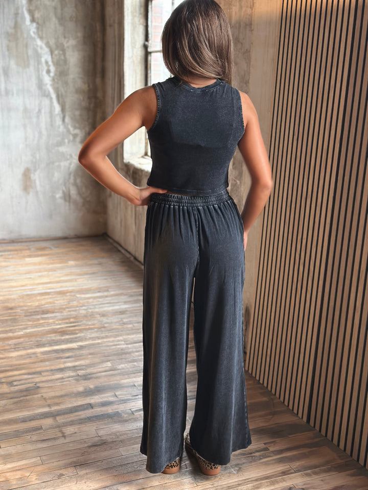 Person wearing a black sleeveless top and wide-leg pants standing in a room with wooden flooring and a textured wall.