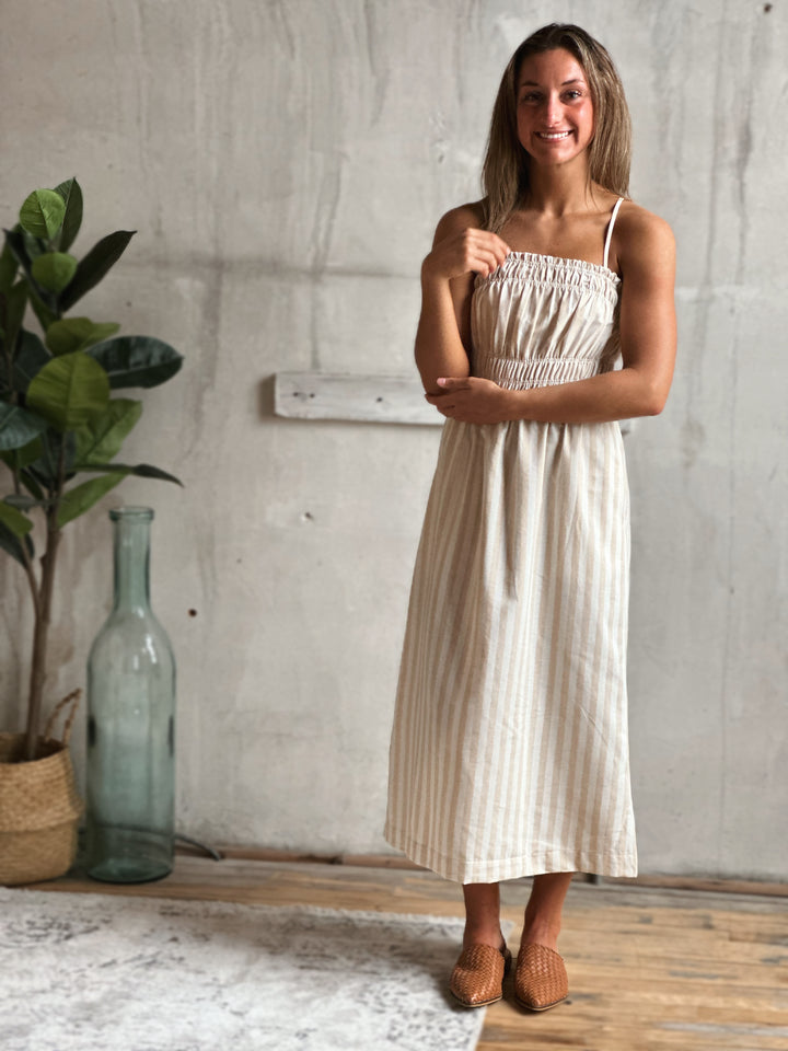 Woman wearing a white and cream striped dress standing in a room with a plant and a bottle.