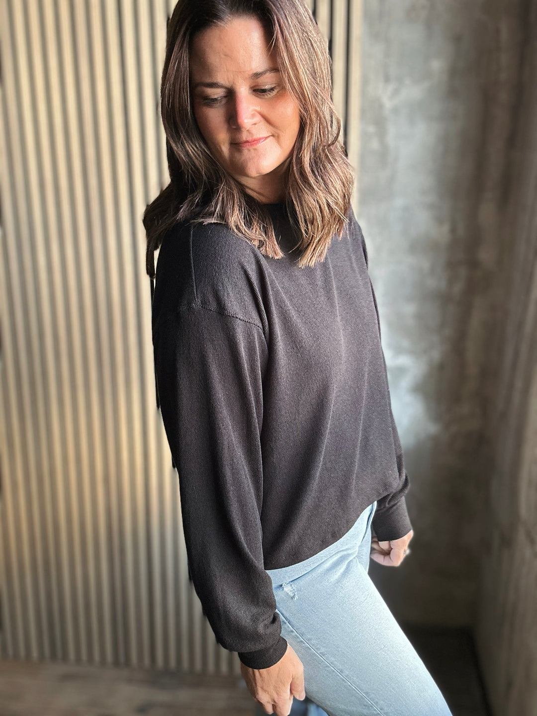 Woman wearing a dark gray sweater and light blue jeans against a textured wall.