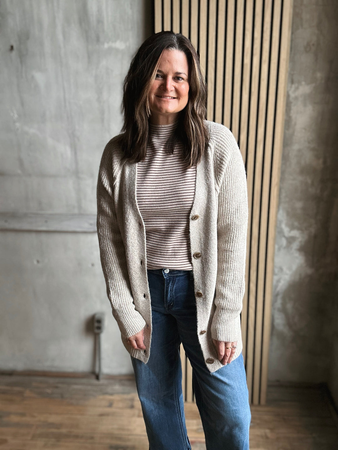 Woman wearing a beige cardigan over a striped shirt with blue jeans, standing against a concrete wall.