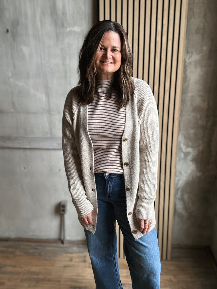 Woman wearing a beige cardigan over a striped shirt with blue jeans, standing against a concrete wall.