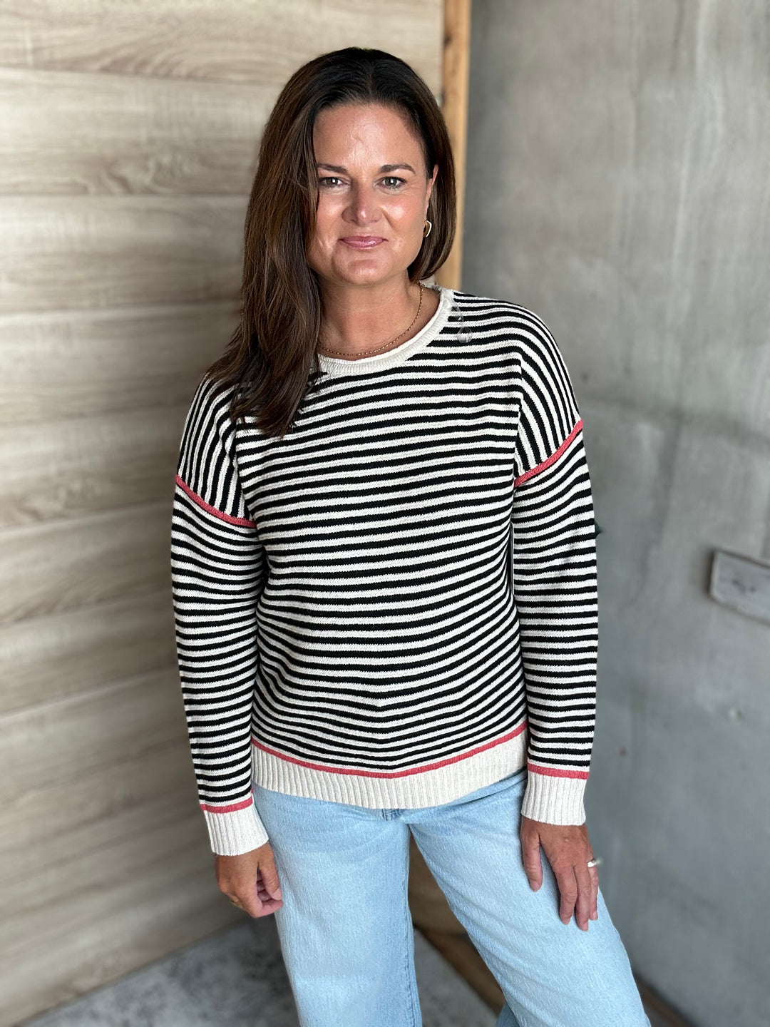 A woman wearing a black and white horizontally striped knit sweater with thin pink accent stripes on the shoulders and cuffs, paired with light blue jeans, standing in front of a wood-paneled wall.