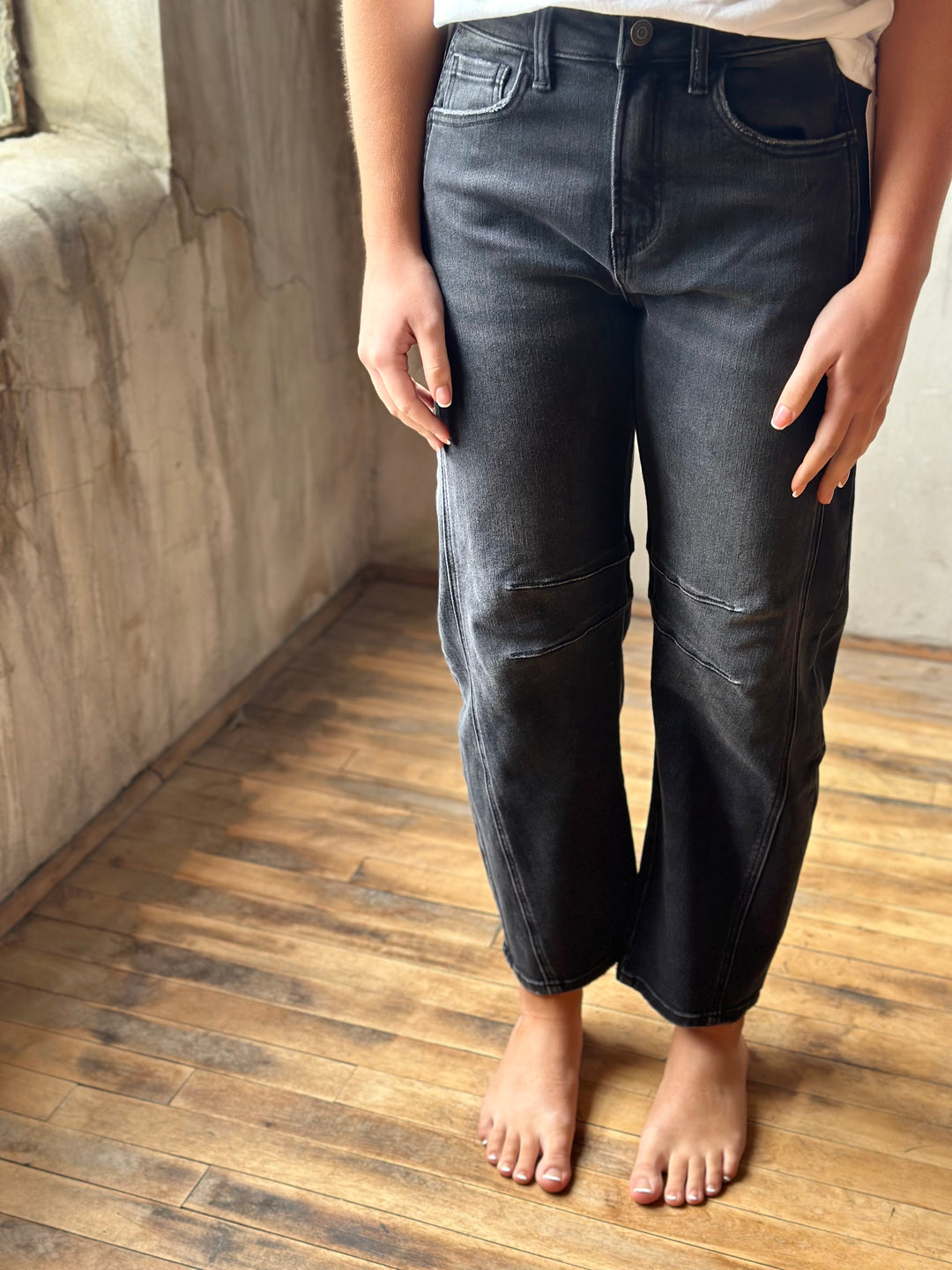 A person wearing dark gray washed denim barrel jeans standing barefoot on a wooden floor against a textured concrete wall.