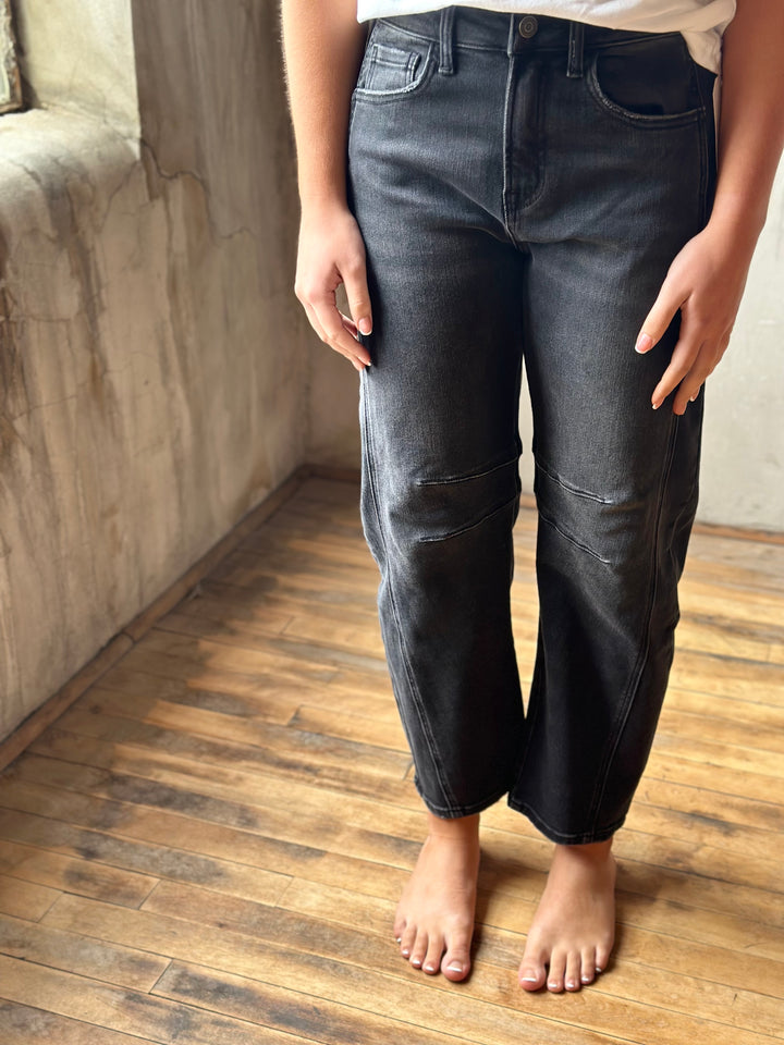 A person wearing dark gray washed denim barrel jeans standing barefoot on a wooden floor against a textured concrete wall.