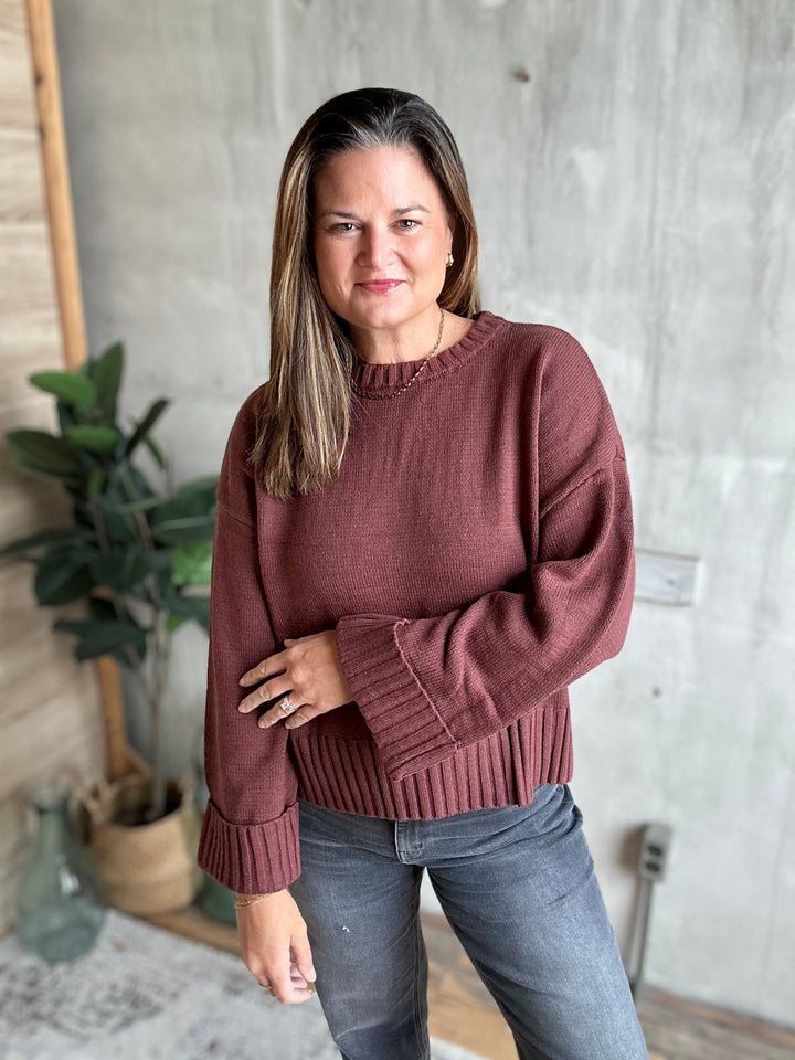 A woman wearing a ribbed crewneck sweater in a dusty rose color, paired with gray jeans, standing in front of a concrete wall with a potted plant in the background.