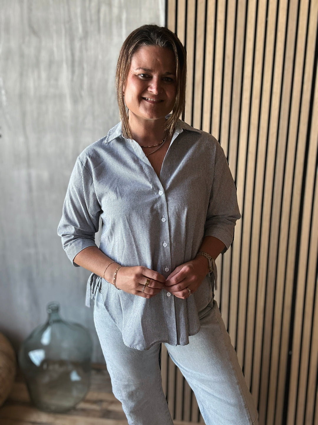 Woman wearing a light gray stripe shirt and pants standing against a wooden paneled wall.