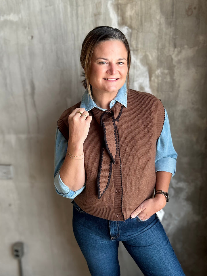 A woman wearing a brown ribbed knit vest over a light blue button-up shirt and blue jeans, standing against a textured concrete wall.