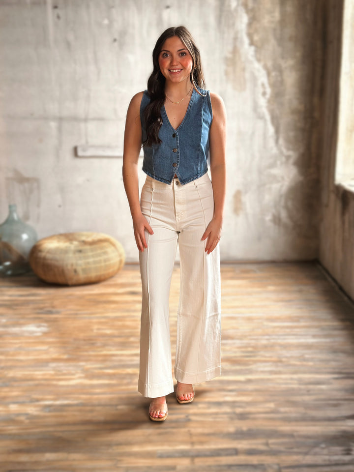 Woman wearing a denim vest and white jeans standing in a room with wooden floor and light-colored walls.