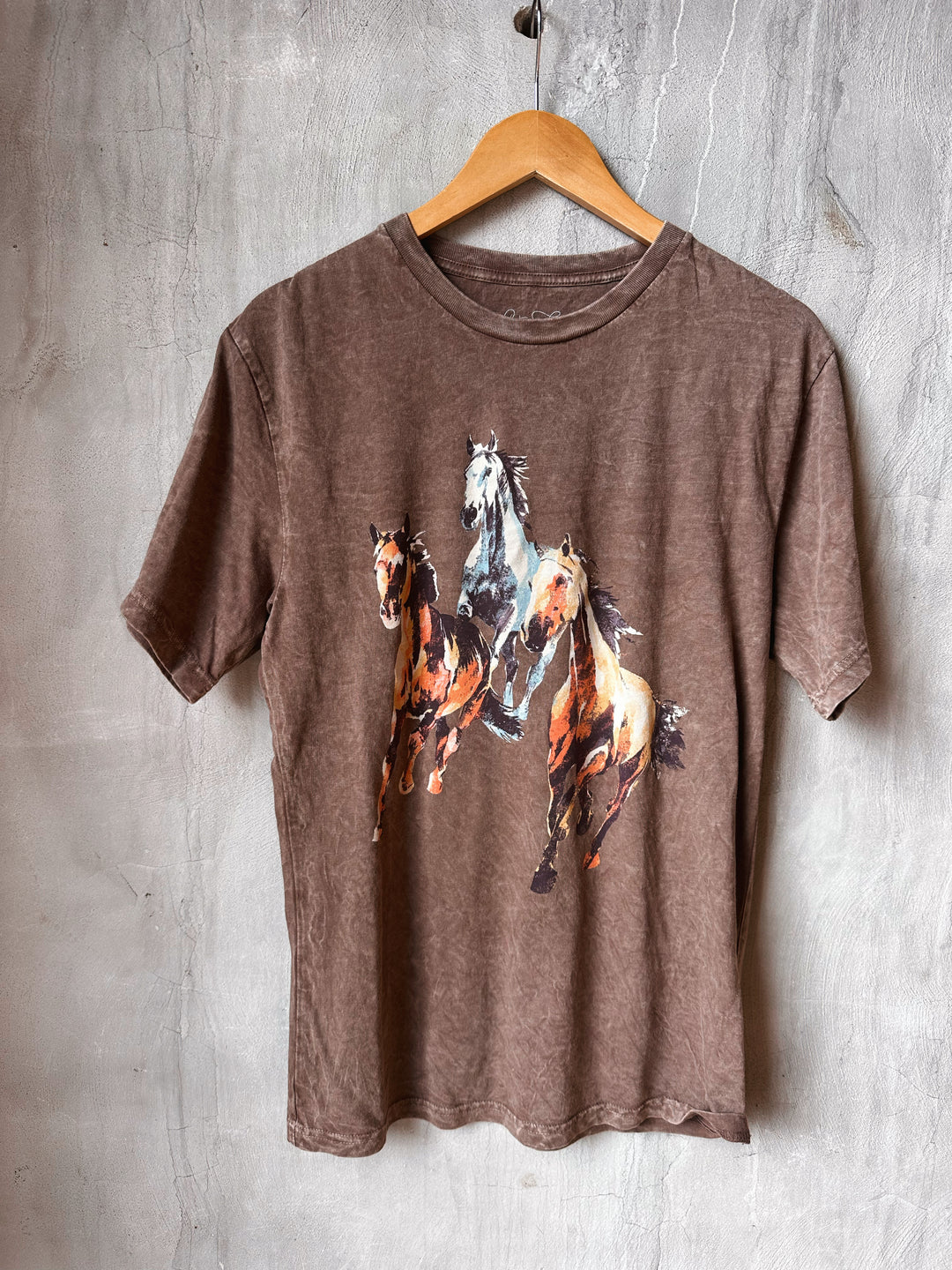 Brown t-shirt with horse design on a wooden hanger against a textured wall.