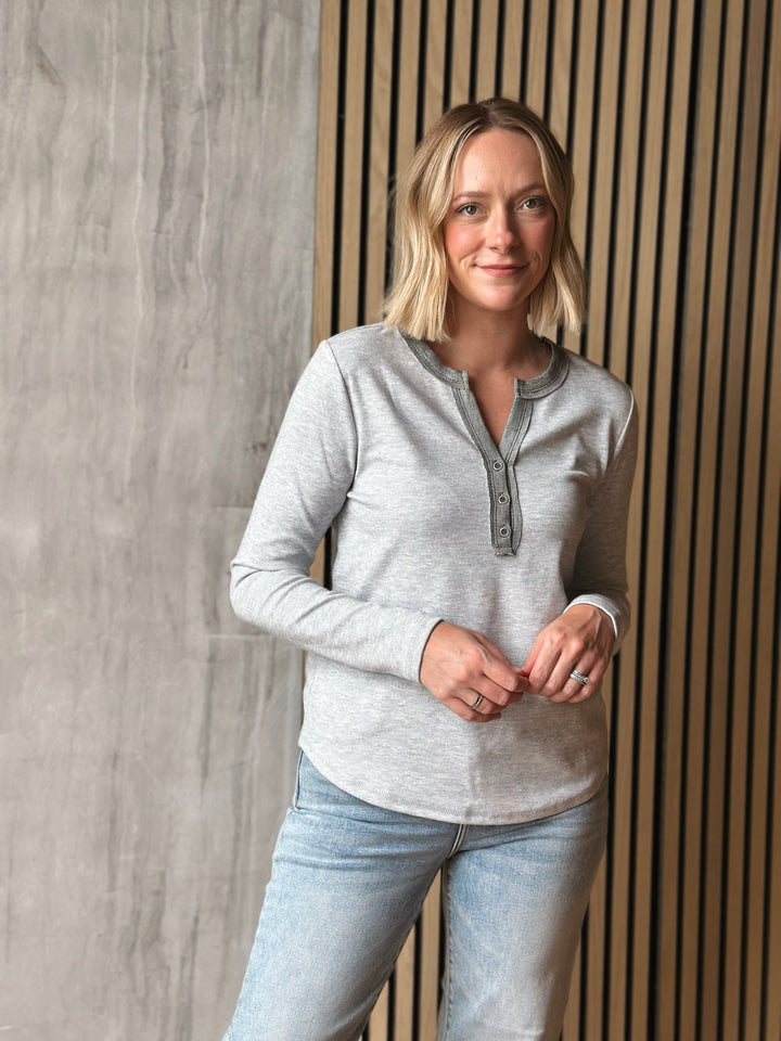 Woman wearing a gray long-sleeve shirt and light blue jeans against a wooden paneled wall.