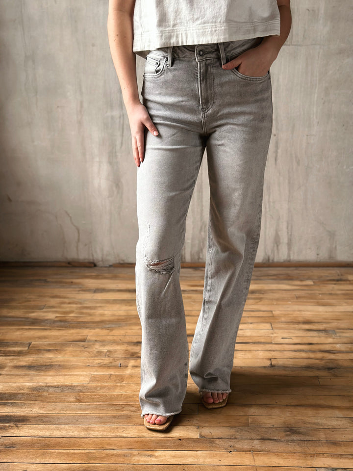 Person wearing light gray jeans standing against a concrete wall on a wooden floor.