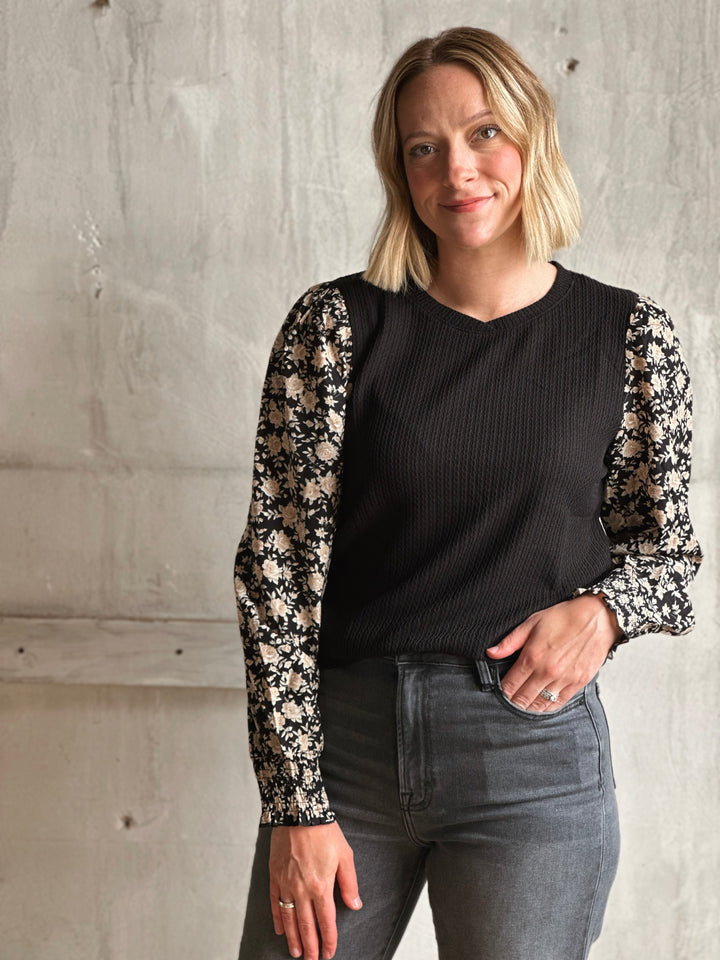 Woman wearing a black top with floral sleeves against a concrete wall.