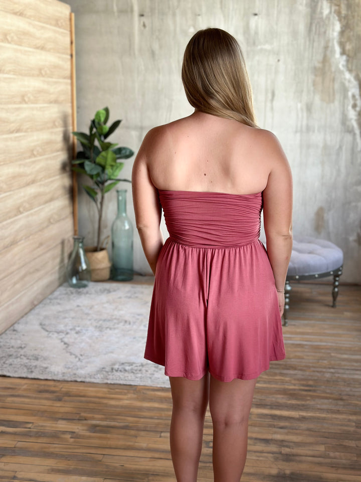 Woman wearing a strapless pink romper in a room with wooden floor and white wall.