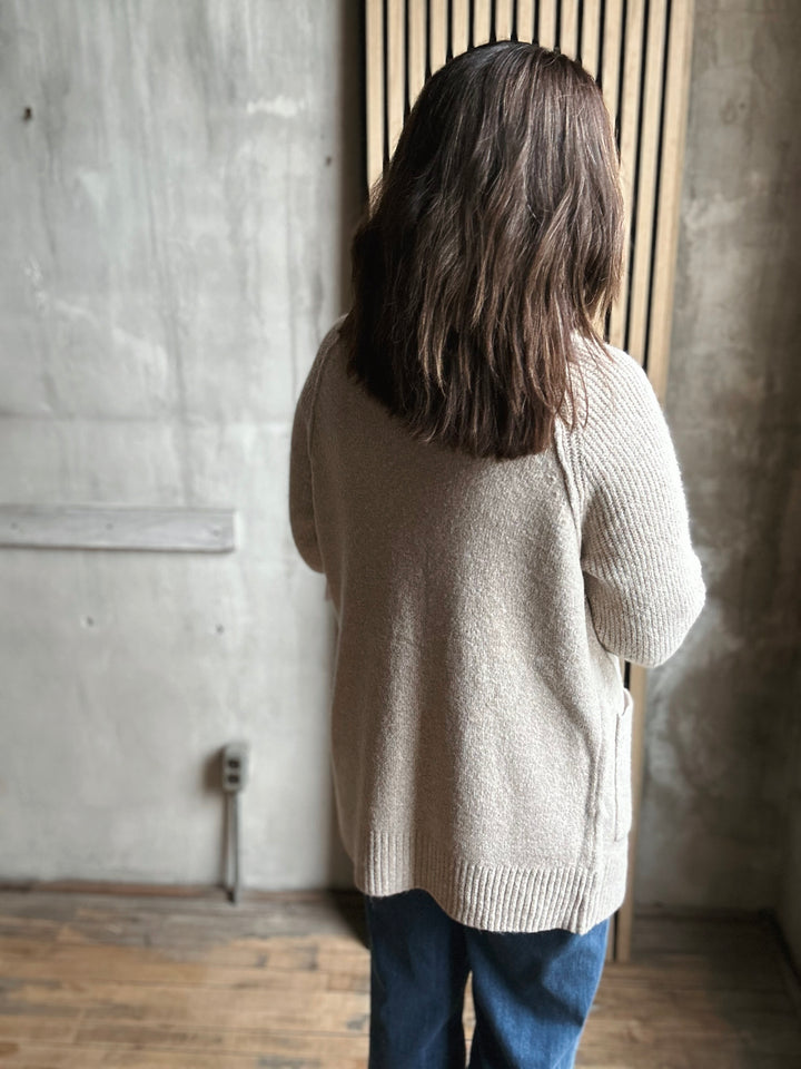 Person wearing a beige sweater and blue jeans standing against a textured wall.