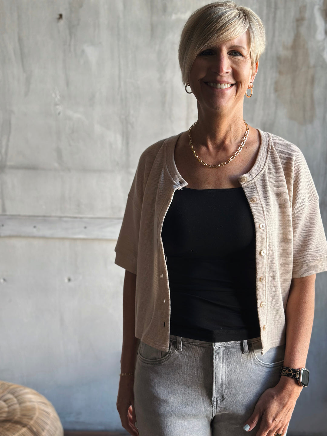 Woman wearing a beige cardigan over a black top and jeans, standing against a concrete wall.