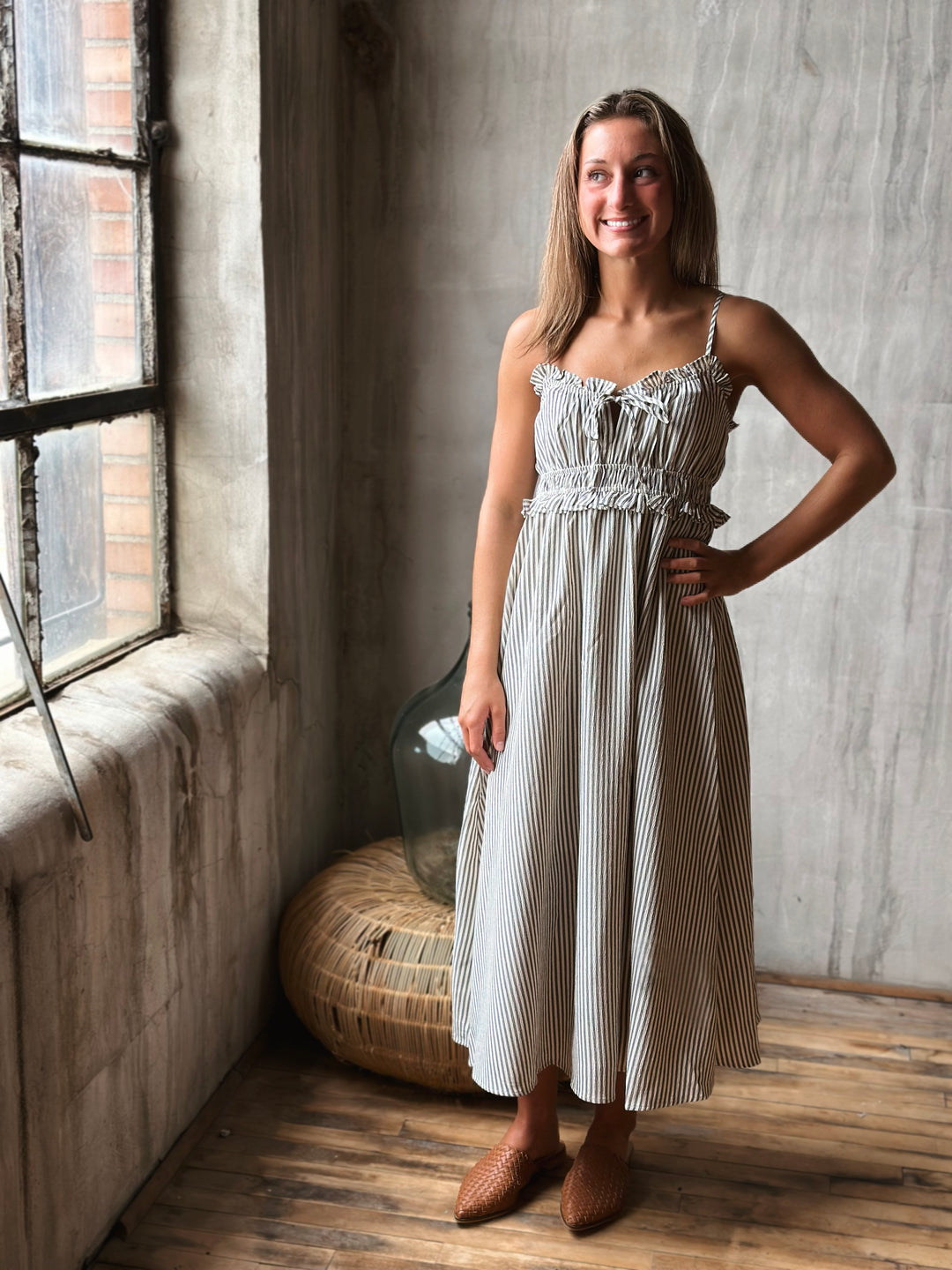 Woman in a white and gray striped dress standing in a room with a window and wooden floor.
