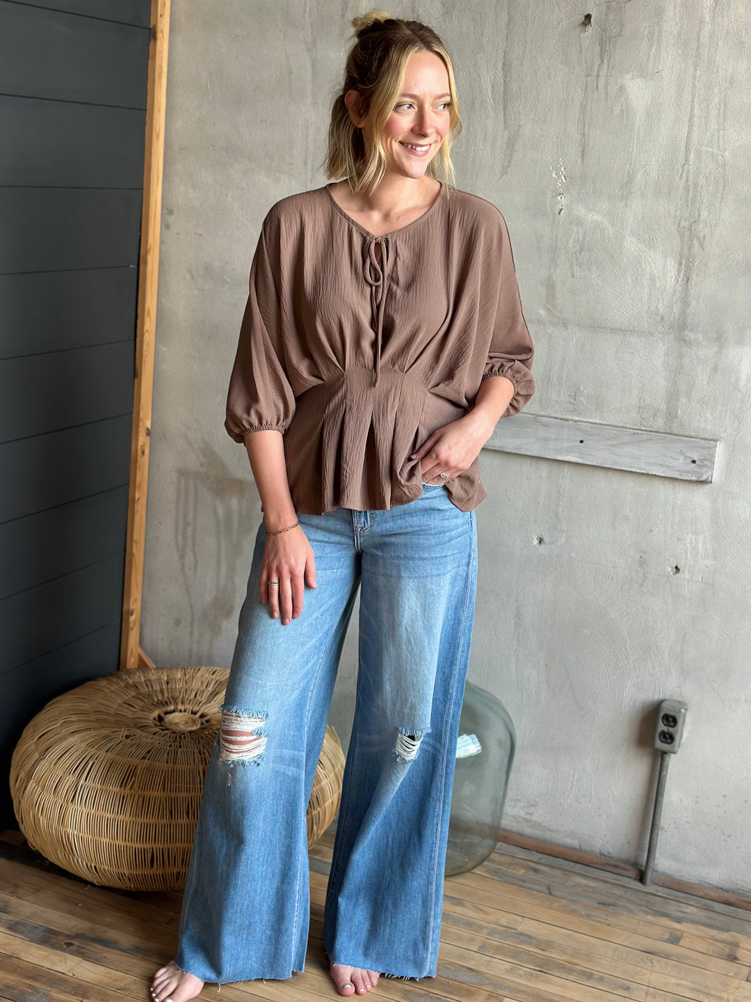 Woman wearing a brown blouse and blue jeans standing in a room with a concrete wall and wooden floor.