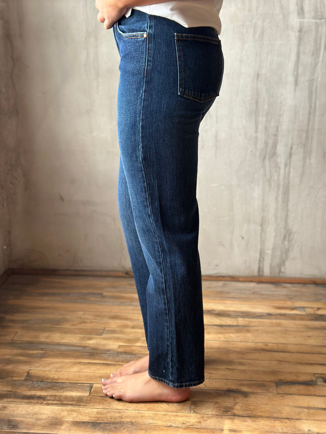 A person shown from the waist down wearing dark blue straight-leg jeans, standing barefoot on a wooden floor against a textured concrete wall.
