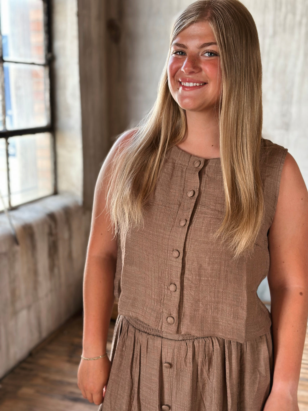 Woman wearing a brown sleeveless top and skirt with buttons standing indoors with a window in the background.