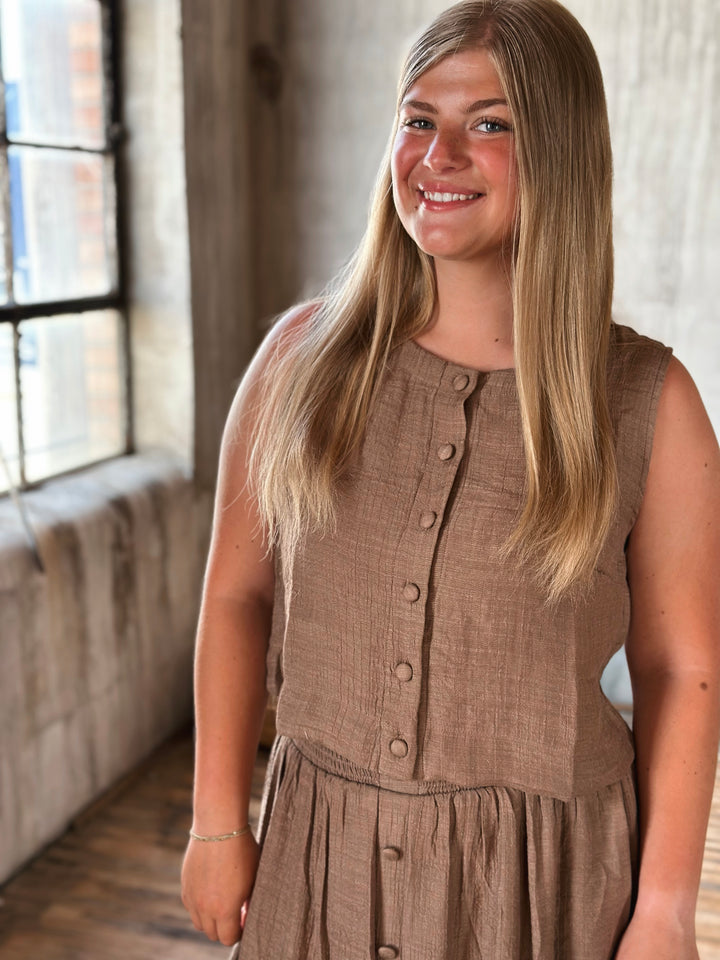 Woman wearing a brown sleeveless top and skirt with buttons standing indoors with a window in the background.