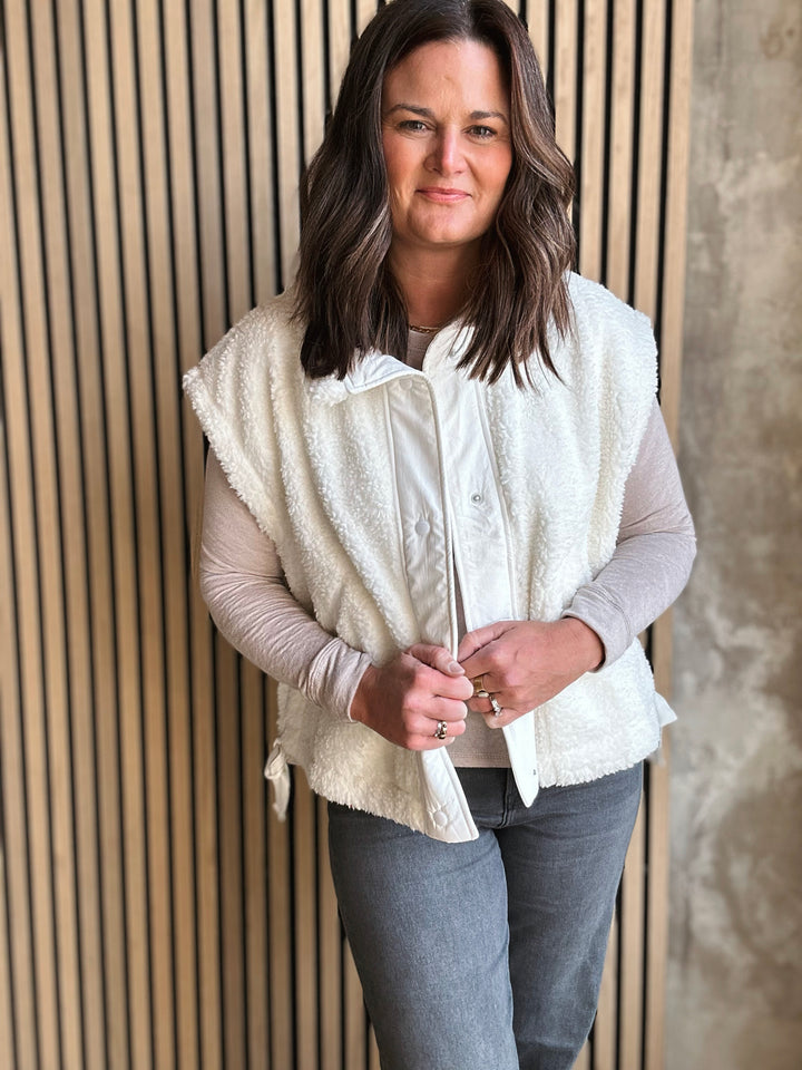 Woman wearing a white vest over a light pink shirt and gray pants against a wooden paneled wall.