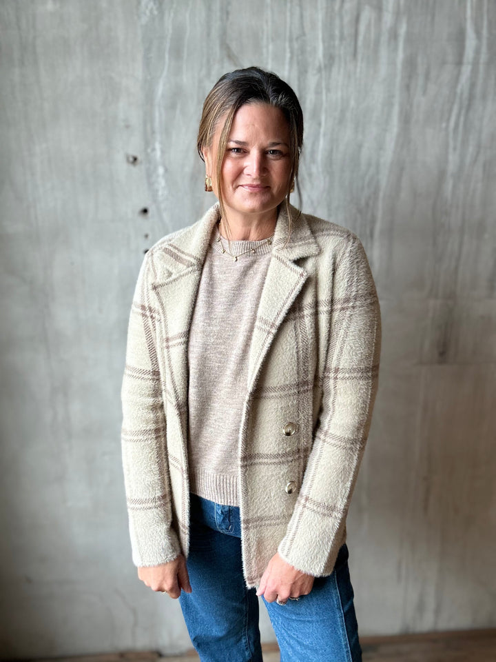 A woman wearing a beige and brown plaid button-front cardigan over a beige knit sweater and blue jeans, standing against a textured gray concrete wall.