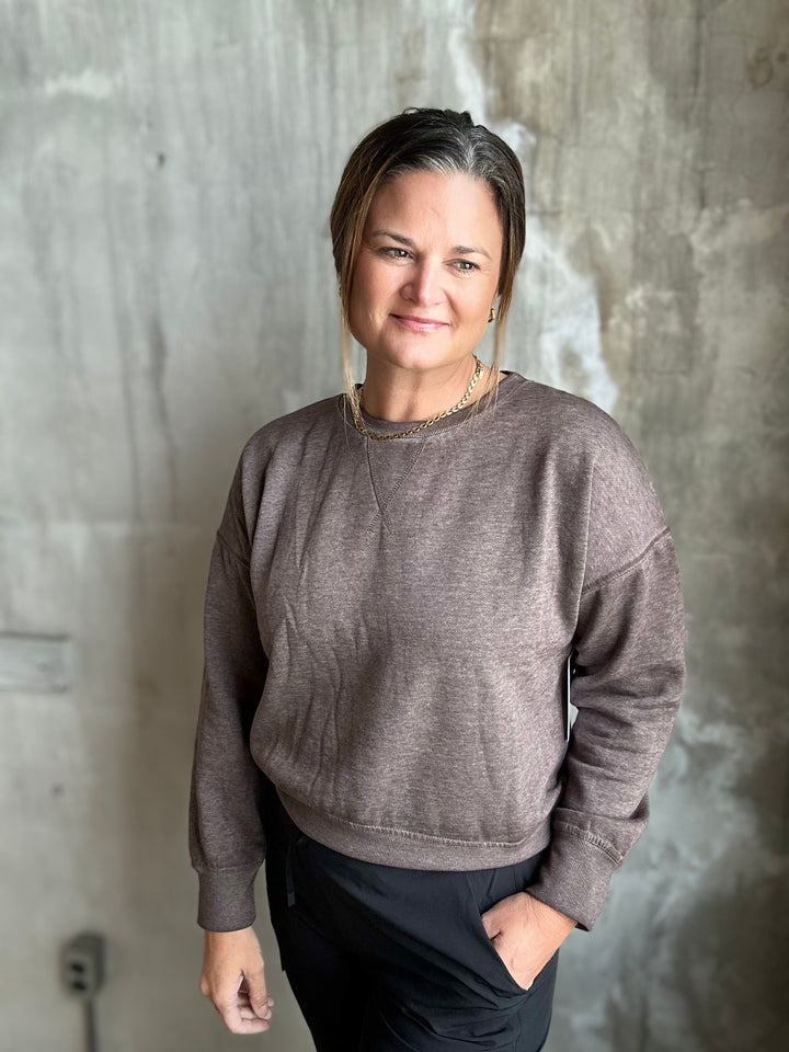 A woman wearing a heathered taupe-brown crewneck sweatshirt and black pants, standing against a textured concrete wall.