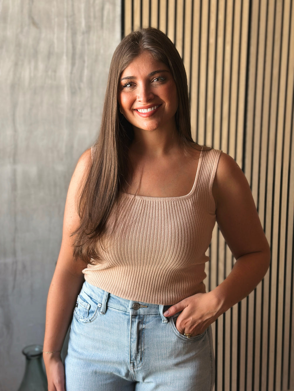 Woman wearing a beige ribbed tank top and light blue jeans against a wooden paneled wall.