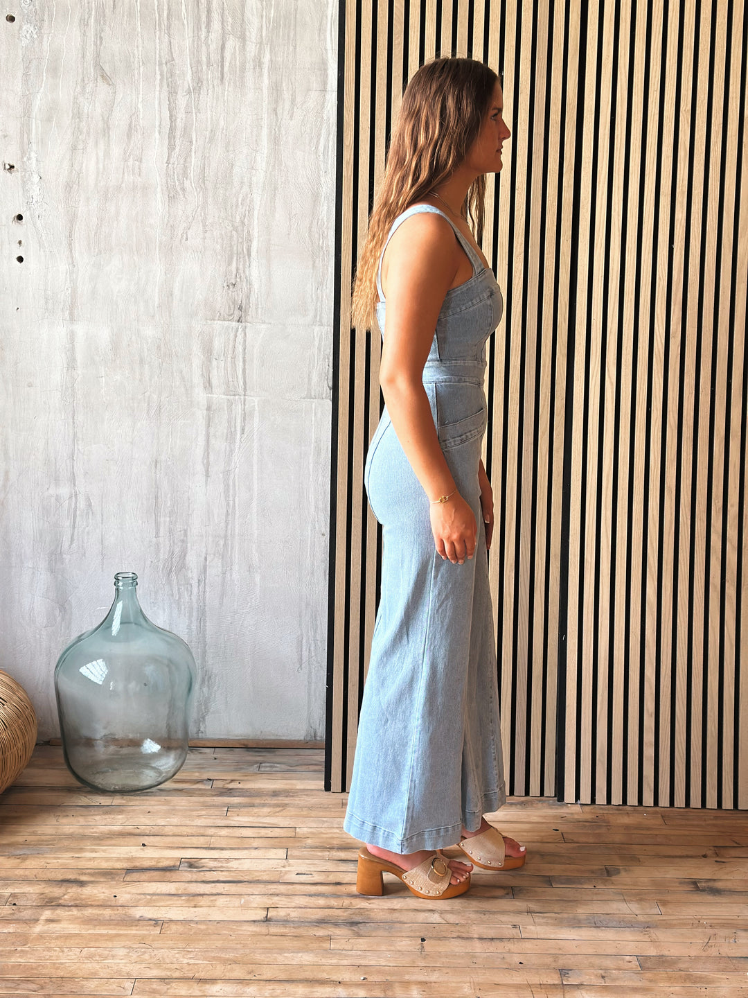 A woman in a light blue denim jumpsuit standing in a room with a wooden floor and striped wall.