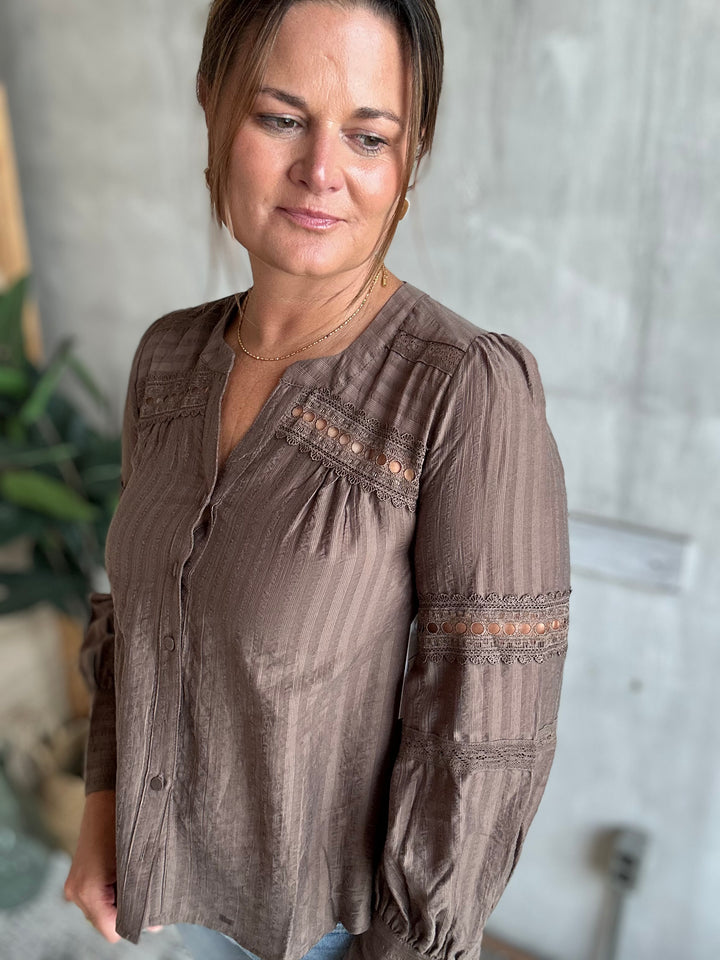 A woman wearing a brown long-sleeve blouse with vertical stripes and lace-up embroidery details on the chest and sleeves, standing against a textured gray wall.