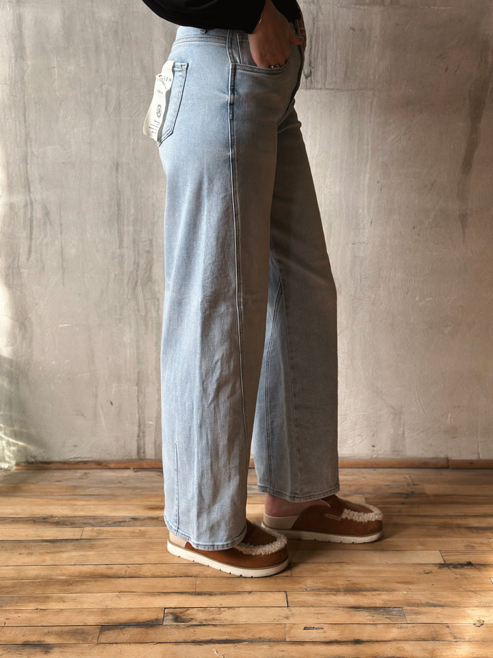 Person wearing light blue wide-leg jeans and brown shoes against a plain wall.