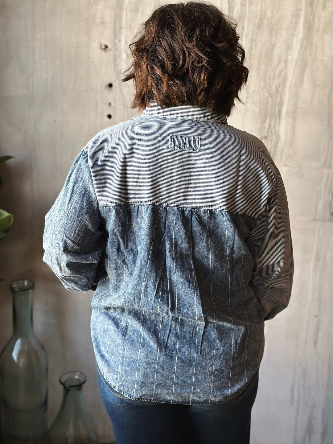 Person wearing a blue denim shirt with a brand logo, standing in front of a textured wall.