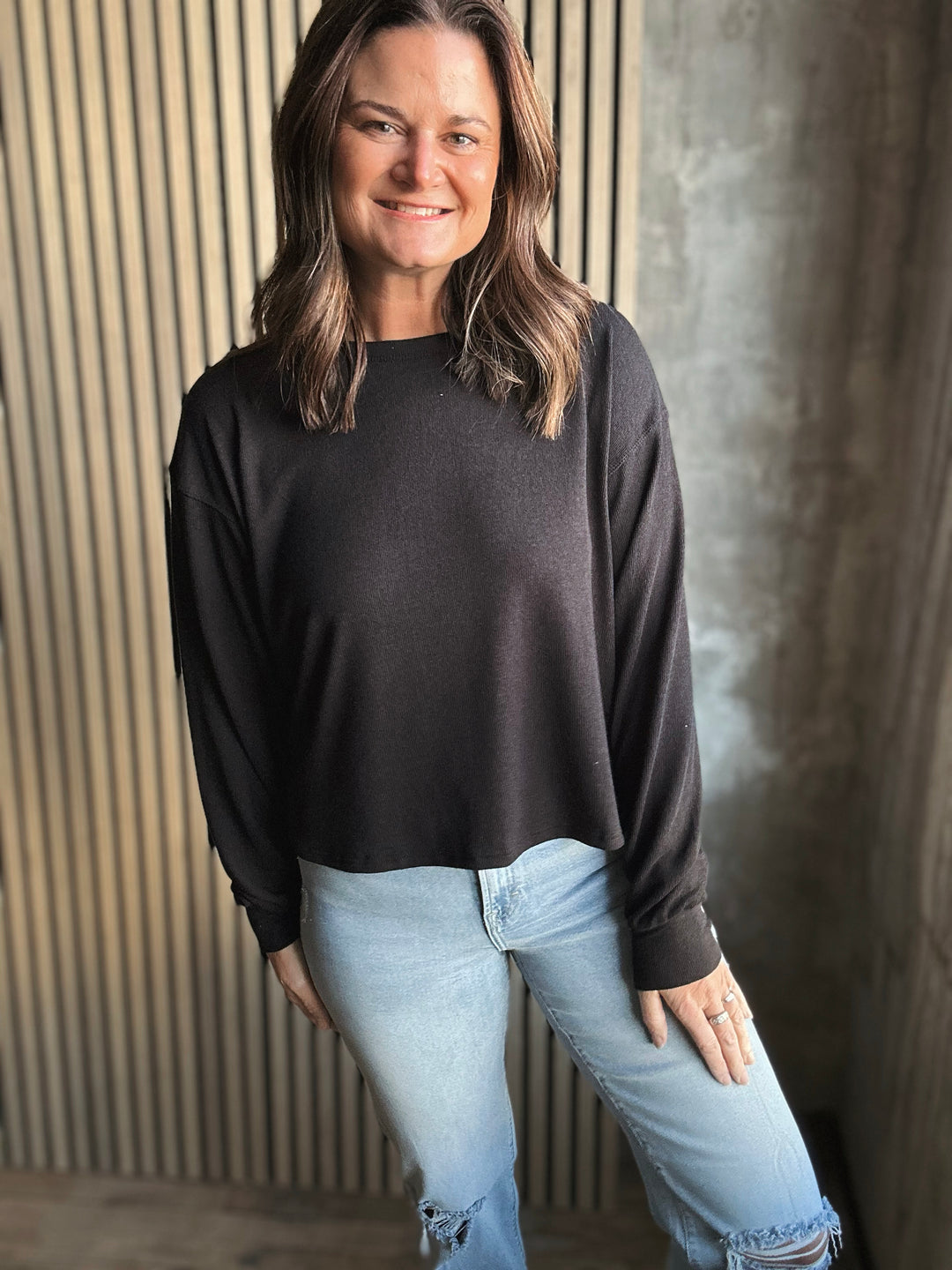 Woman wearing a black sweater and light blue jeans against a textured wall.