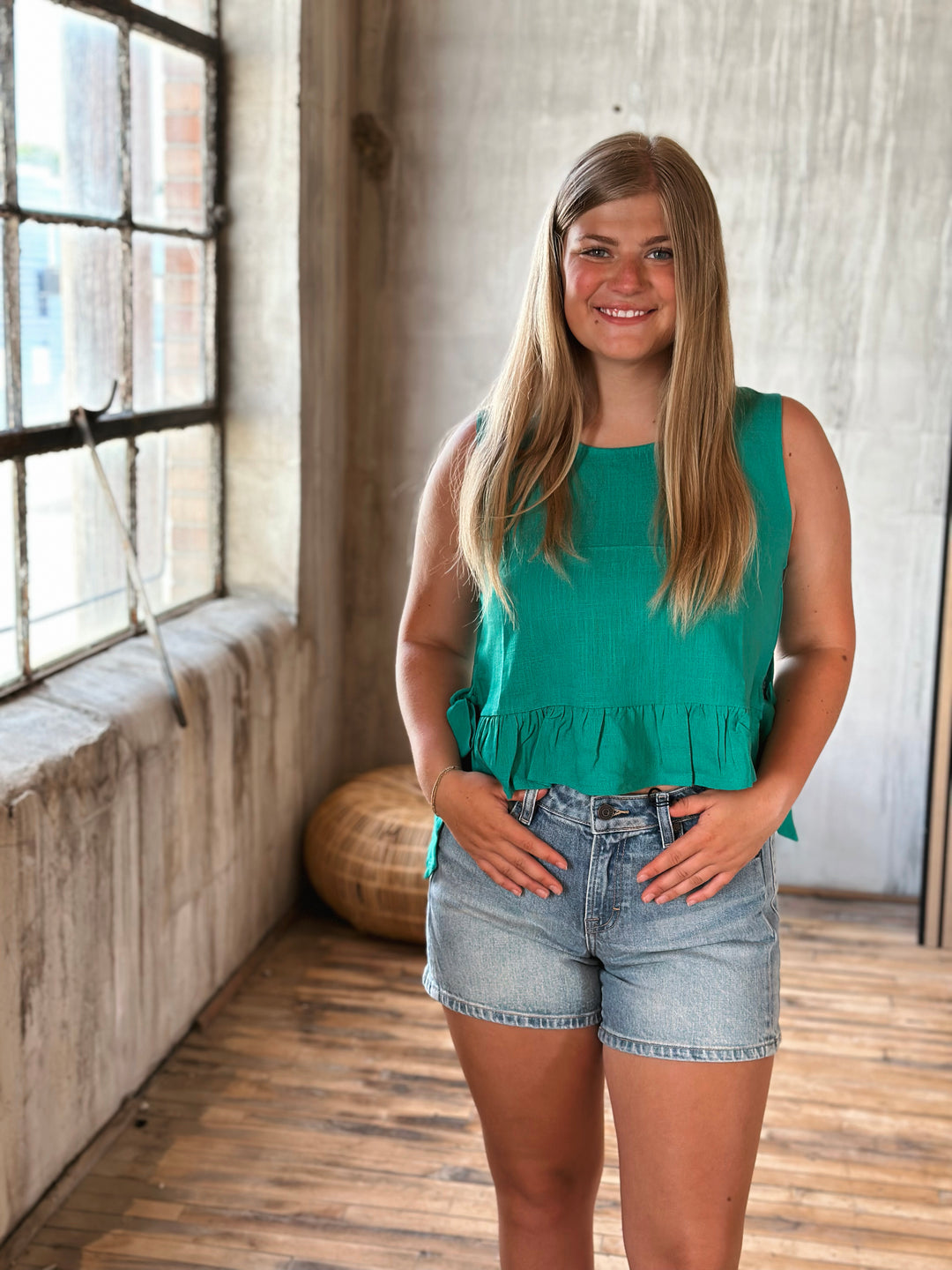 Woman in a green sleeveless top with a ruffle at the bottom and denim shorts standing in a room with wooden floor and large window.