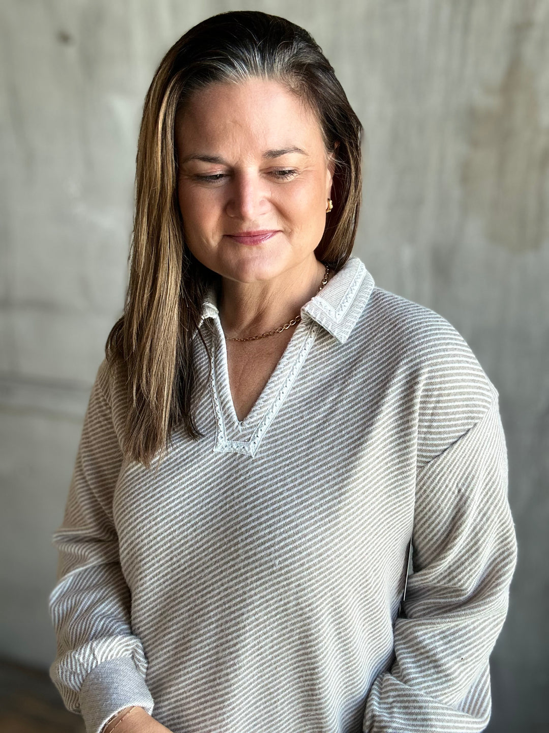 A woman with shoulder-length brown hair with gray streaks, wearing a light gray and white vertically striped long-sleeve polo shirt, looking down with a soft expression against a textured gray wall.
