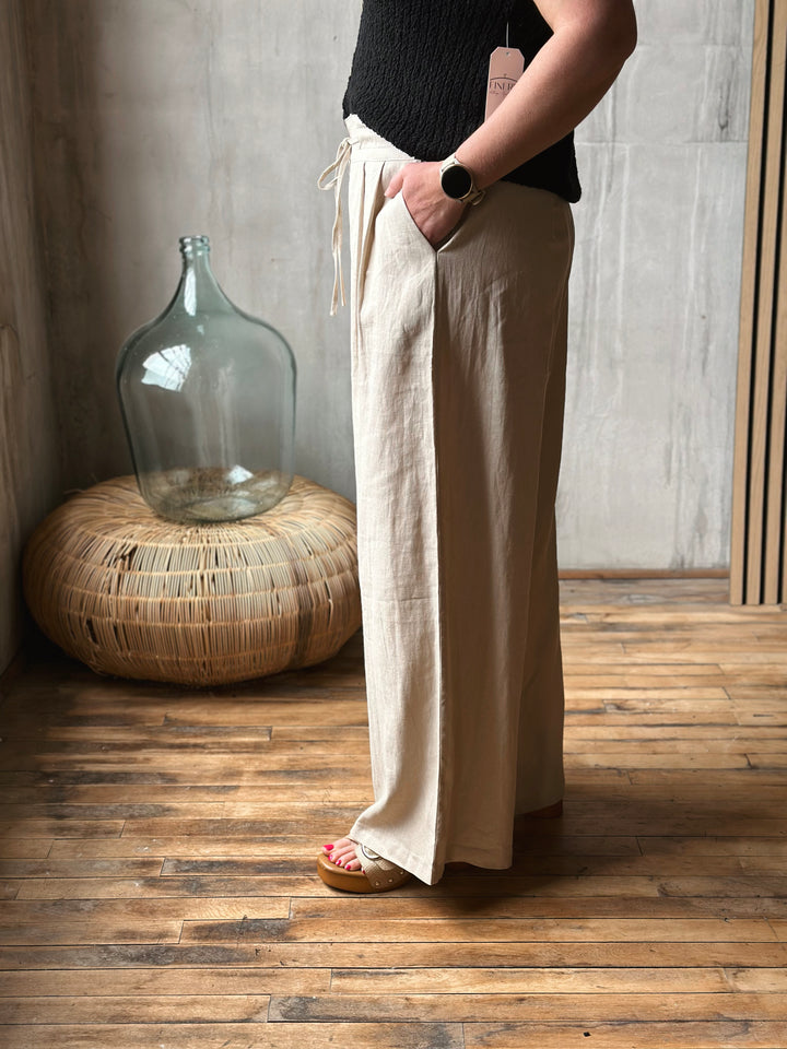 Person wearing beige wide-leg pants standing in a room with wooden floor and gray wall.