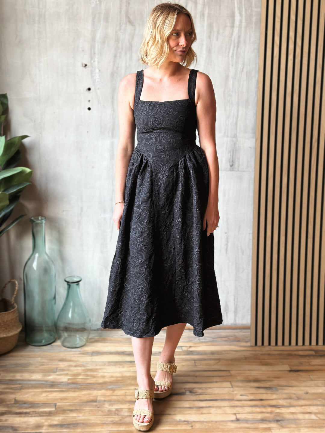 Woman wearing a black sleeveless dress standing indoors with decorative vases and plants in the background.