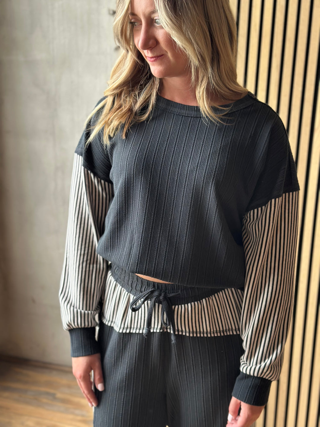 A woman wearing a dark charcoal ribbed long-sleeve top with beige and black striped sleeves, paired with matching dark ribbed pants, standing indoors against a textured wall and slatted wood paneling.