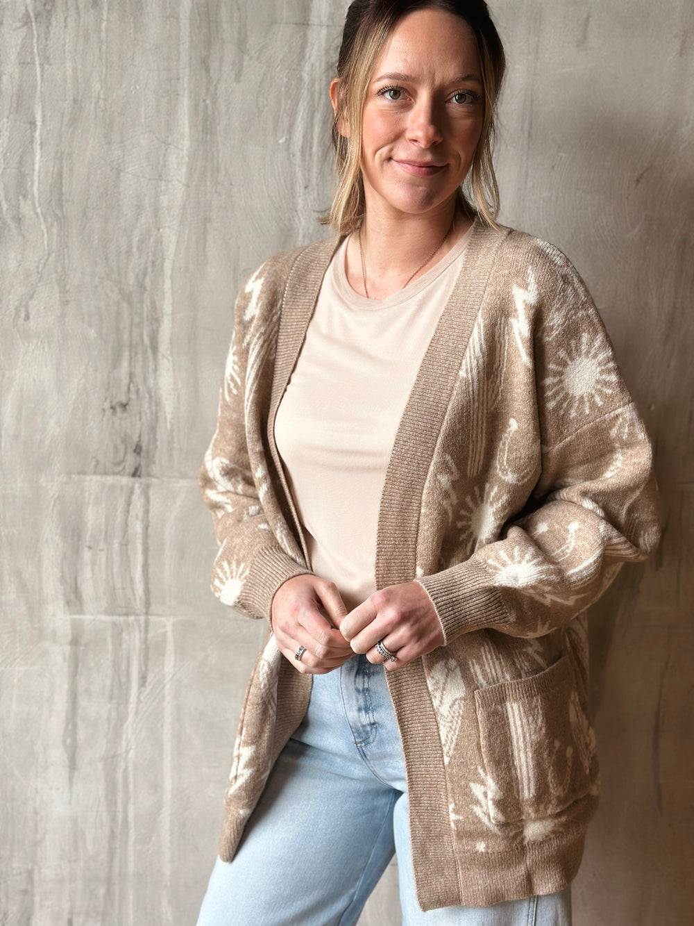 Woman wearing a beige patterned cardigan over a white shirt and blue jeans against a gray concrete wall.