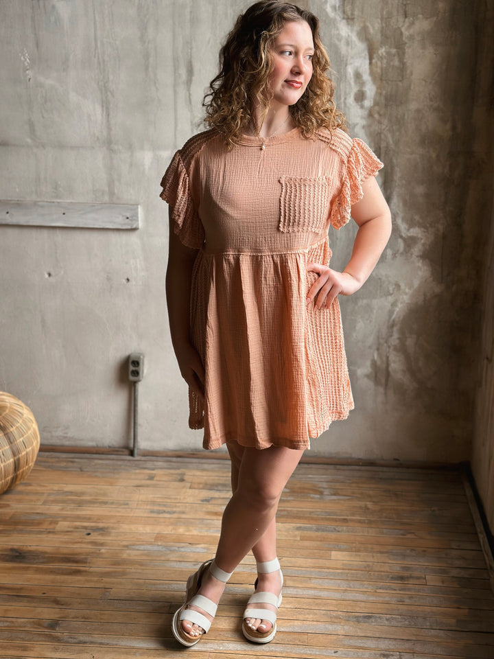 Woman wearing a peach dress standing in a room with a wooden floor and concrete wall.