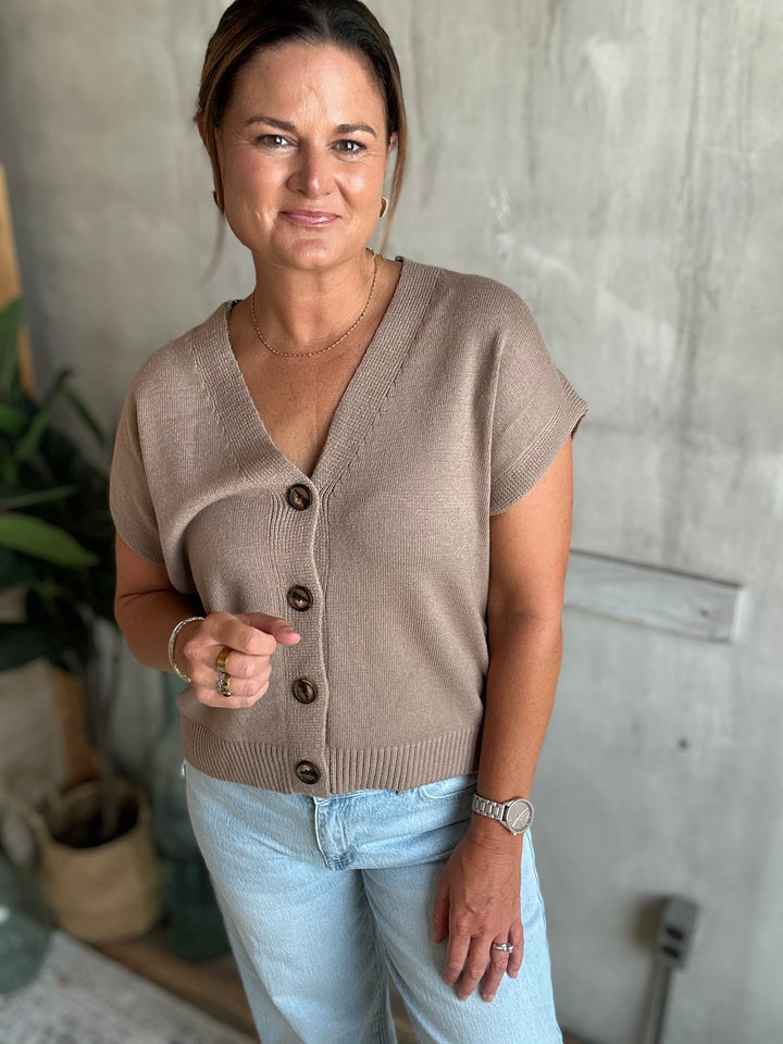 A woman wearing a taupe short-sleeve knit cardigan with brown buttons and light blue jeans, standing against a textured gray concrete wall.