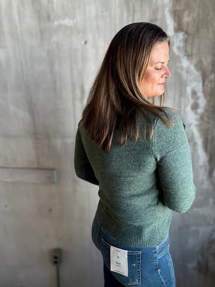 A woman with long brown hair is shown from the back and side, wearing a sage green knit sweater and blue jeans against a textured concrete wall.
